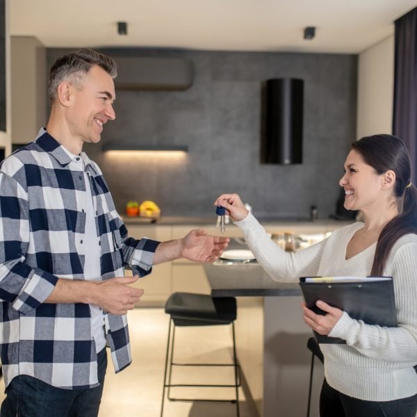 New housing. Happy middle aged man in casual clothes taking keys from welcoming smiling pretty female realtor standing opposite each other sideways to camera in room
