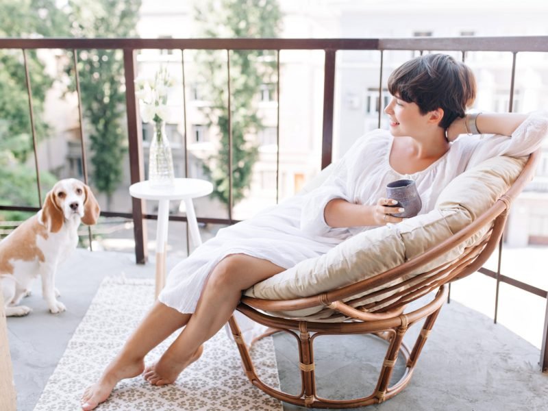 Relaxed barefooted girl in white dress sitting in chair on balcony and holding cup of tea. Adorable brunette woman drinking coffee on terrace and looking with smile at beagle dog.